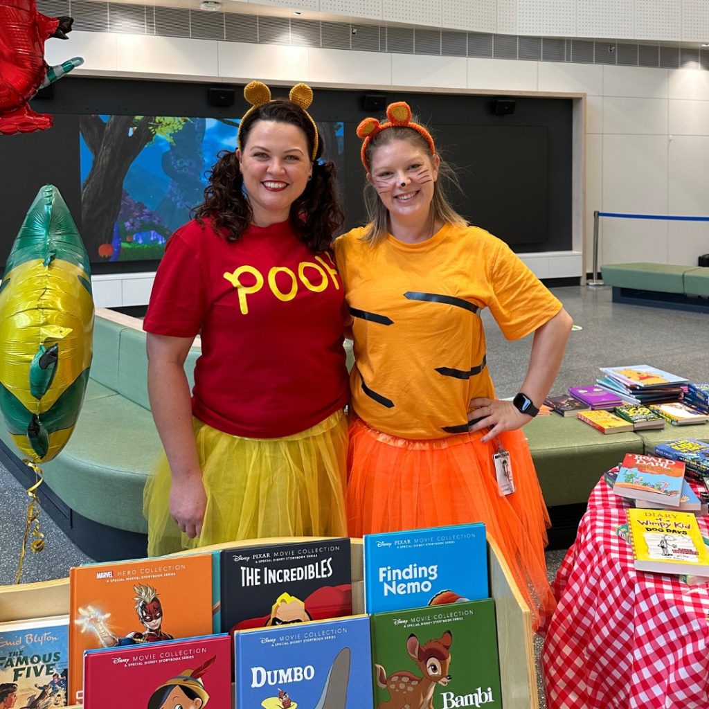 Two women dressed as Winnie-the-Pooh and Tigger the tiger smile for a photo beside a book display.